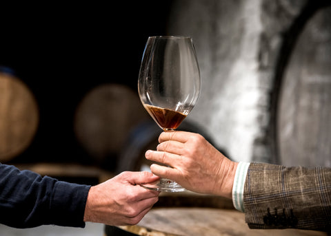 Side view of hands holding a wine glass during a tasting, with aged wine bottles visible in the background